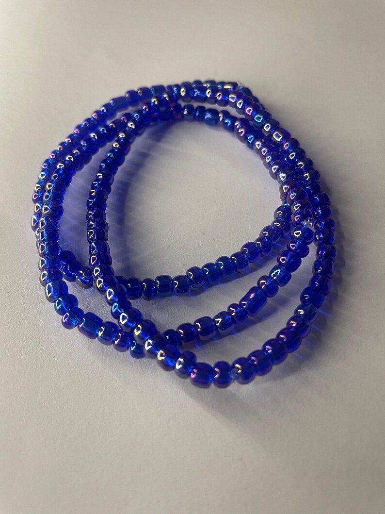 K&ouml;nnte beinhalten: Eine blaue Perlenkette mit schillernden Perlen. Die Kette besteht aus mehreren Perlenstr&auml;ngen und ist auf einer wei&szlig;en Oberfl&auml;che aufgerollt.