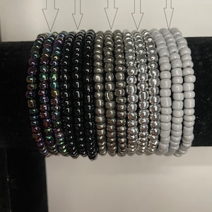 May include: A collection of beaded bracelets in various colors, including black chrome, black, metallic silver, silver, and gray. The bracelets are stacked together, showcasing the different shades and finishes of the beads.