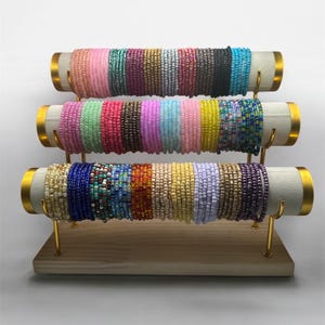 May include: A three-tiered display stand showcasing numerous beaded bracelets. The bracelets are arranged in a rainbow of colors, including pink, green, red, purple, yellow, and blue. The stand has a wooden base with gold-colored supports and white cylindrical holders.