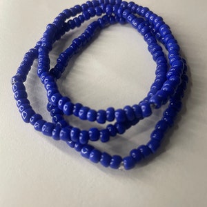 K&ouml;nnte beinhalten: Ein blaues Perlenarmband. Das Armband besteht aus kleinen, runden Perlen, die auf einer d&uuml;nnen Schnur aufgef&auml;delt sind. Das Armband ist auf einer wei&szlig;en Oberfl&auml;che aufgerollt.
