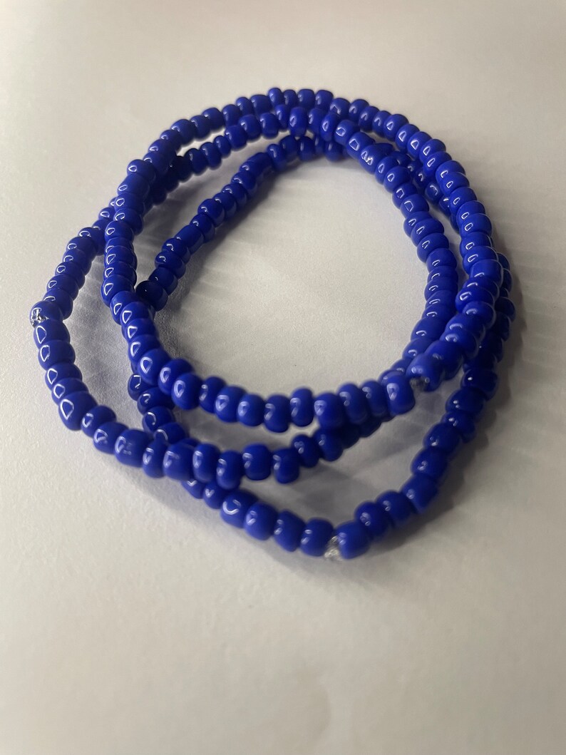 K&ouml;nnte beinhalten: Ein blaues Perlenarmband. Das Armband besteht aus kleinen, runden Perlen, die auf einer d&uuml;nnen Schnur aufgef&auml;delt sind. Das Armband ist auf einer wei&szlig;en Oberfl&auml;che aufgerollt.
