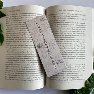 May include: A white bookmark with a floral design and the text "You are the author of your story." The bookmark is lying on top of an open book.