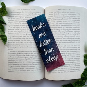 Könnte beinhalten: Ein Lesezeichen im Galaxie-Design mit dem Text "books are better than sleep" in weißen Buchstaben. Das Lesezeichen ist aus Papier und hat einen Farbverlauf von Blau, Lila und Rosa.