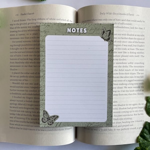 Könnte beinhalten: Ein grüner und weißer Notizblock mit dem Wort "NOTES" in Schwarz oben. Der Notizblock ist mit einem Schmetterlingsdesign auf der linken und rechten Seite verziert. Der Notizblock liegt auf einem geöffneten Buch.