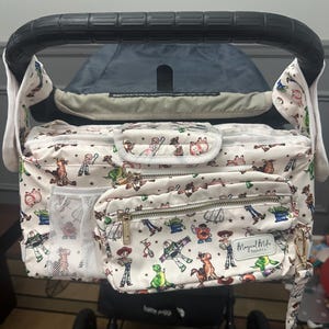 May include: A white stroller organizer bag with a colorful Toy Story character print. The bag has multiple zippered compartments, a mesh pocket, and a gold-colored zipper. The bag is attached to a black stroller handle.