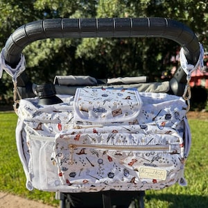 Puede incluir: Un organizador de cochecito blanco con un estampado de temática de Harry Potter. El organizador tiene un bolsillo con cremallera, un bolsillo de malla y un portavasos. El estampado presenta estrellas, varitas, búhos y otros símbolos de Harry Potter. El texto "Magical Misfits" está impreso en el organizador.