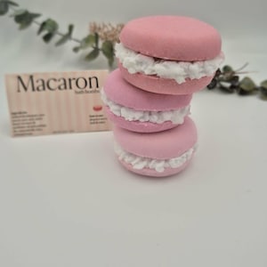 Peut inclure: Trois bombes de bain roses en forme de macarons avec une garniture blanche sont empilées. Une boîte avec le mot "Macaron" est en arrière-plan. Les bombes de bain sont conçues pour ressembler à la pâtisserie française.
