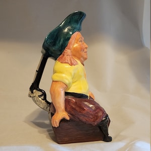 May include: A ceramic pitcher shaped like a pirate sitting on a wooden box. The pirate is wearing a green hat, a yellow shirt, brown pants, and black boots. The pitcher has a handle shaped like a gun.