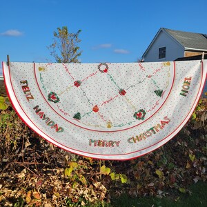 May include: White tablecloth with red trim and a festive Christmas design. It features red and green polka dots, a wreath, presents, and the words "Feliz Navidad", "Merry Christmas", and "Noel".