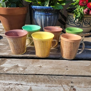 May include: A collection of seven vintage coffee mugs with a woven texture. The mugs have clear handles and are in shades of pink, green, yellow, and peach. They are arranged on a weathered wooden surface.