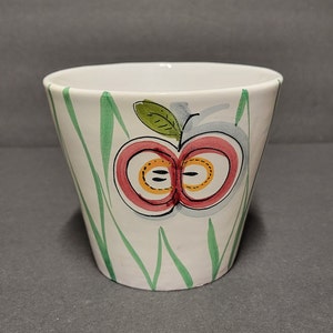 May include: White ceramic planter with a green, red, and yellow apple design. The apple is cut in half and has a green leaf.