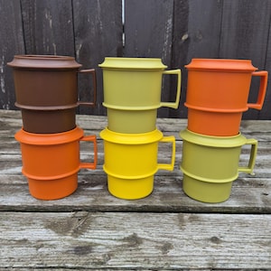 May include: A collection of six vintage plastic mugs in various colors, including brown, orange, yellow, and green. Each mug has a handle and a lid, stacked in pairs. The mugs are arranged on a weathered wooden surface.
