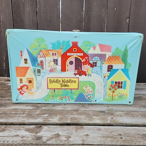 May include: A vintage, light blue toy carrying case with a colorful illustration of a town scene. The illustration includes houses, a fire station, and the text "Liddle Kiddles Town". The case has a metal clasp and is likely from the 1960s or 1970s.