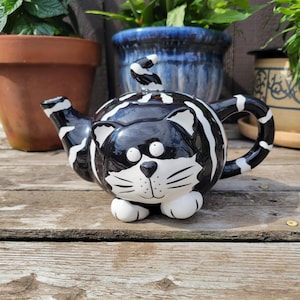 Könnte beinhalten: Eine skurrile, schwarz-weiß geformte Katze-Teekanne. Die Keramik-Teekanne hat eine glänzende Oberfläche mit schwarzen Streifen und einem Katzengesicht-Design. Der Griff und der Ausguss sind ebenfalls schwarz-weiß gestreift. Die Teekanne steht auf einer Holzoberfläche.