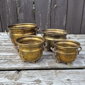 May include: A set of three brass pots with rounded bodies, each featuring two side handles and three small feet. The pots have a polished metallic finish with visible signs of aging and oxidation, showing varying sizes in a nested arrangement.