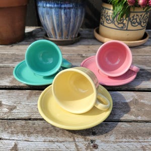 Op de afbeelding: Drie kleurrijke keramische theekopjes en schotels in teal, roze en geel zijn gerangschikt op een verweerd houten oppervlak. De kopjes zijn rond met oren en de schotels zijn cirkelvormig. Op de achtergrond staan potplanten.
