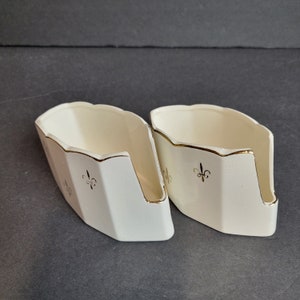 May include: Two white ceramic napkin holders with gold trim and a fleur-de-lis design.