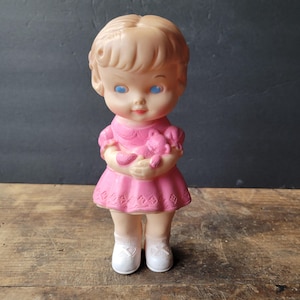 Peut inclure: Poupée vintage avec des yeux bleus, vêtue d'une robe rose et tenant un ours en peluche rose. La poupée a des chaussettes et des chaussures blanches. Les cheveux de la poupée sont brun clair.