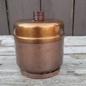 Op de afbeelding: Een koperkleurige metalen container met een deksel. De container heeft een afgeronde vorm en een glad oppervlak. Het deksel heeft een verhoogd, getextureerd ontwerp.