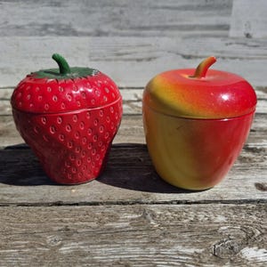 May include: Two ceramic fruit-shaped containers. One is a red strawberry with a green stem, the other is a red and yellow apple with a brown stem. Both containers have lids and sit on a wooden surface.