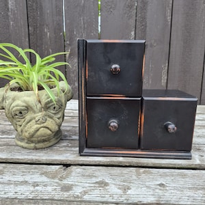 May include: A black wooden storage unit with three drawers arranged in a stepped configuration, featuring small round knobs and distressed edges. Beside it sits a gray stone planter shaped like a pug dog head containing a green spider plant.