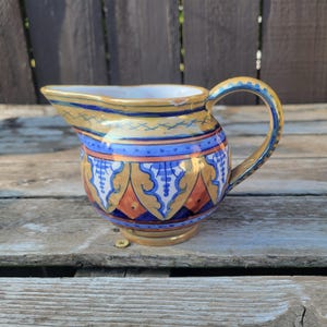 May include: A small, decorative ceramic pitcher with a curved handle and spout. The pitcher features a vibrant design with blue, orange, and gold patterns. The pitcher is sitting on a weathered wooden surface.