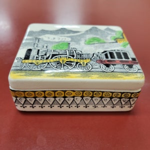 May include: A white ceramic box with a black and white train design on the lid. The train is on a yellow track and there are green hills in the background. The box has a black and white geometric design around the bottom edge.