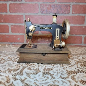 May include: A vintage black and gold sewing machine with the word "SEWING" in gold lettering. The machine sits on a wooden base with a small drawer. The background is a brick wall.