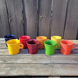 Op de afbeelding: Een verzameling van acht kleurrijke keramische mokken in verschillende tinten, waaronder geel, rood, oranje, groen en blauw. Elke mok heeft een afgeronde body, een klein handvat en een glanzende afwerking. De mokken staan op een verweerd houten oppervlak.