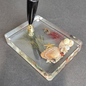 May include: A clear acrylic pen holder with a black pen, seashells, a starfish, and a seahorse inside.