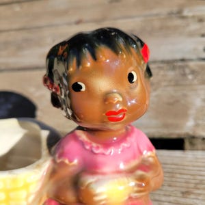 Może przedstawiać: Vintage ceramiczna figurka młodej dziewczyny o ciemnej skórze, czarnych włosach i czerwonych akcentach. Ma na sobie różową sukienkę i czerwone usta. Figurka jest częścią większego elementu dekoracyjnego, prawdopodobnie doniczki lub pojemnika.