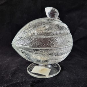 May include: Clear glass decorative container with a textured, leaf-like pattern. The container has a lid with a small, clear knob and sits on a round base. The glass has a reflective quality, and the container is on a dark background.
