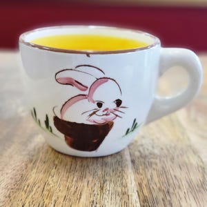 May include: White ceramic teacup with a pink and brown bunny illustration. The cup is filled with a yellow liquid.