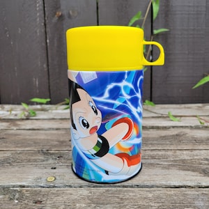 May include: A yellow and blue thermos with a cartoon character on it. The character is a boy with black hair and a red shirt. The thermos has a yellow lid and a handle. The text on the thermos reads "Shogakukan Inc."