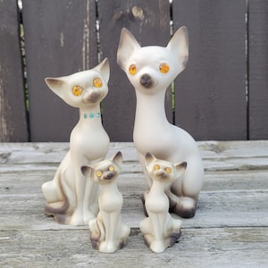Op de afbeelding: Een set van vier keramische Siamese kattenbeeldjes. De katten zijn allemaal wit met zwarte markeringen en hebben oranje ogen. De grootste kat zit rechtop, terwijl de andere drie zitten met hun poten onder zich.