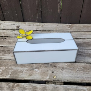 Könnte beinhalten: Eine weiße Taschentuchbox mit einer grauen ovalen Öffnung und einem gelben Schmetterlingsakzent. Die Box hat eine rechteckige Form mit einem grauen Rand und steht auf einer verwitterten Holzoberfläche.