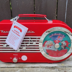 Peut inclure: Radio vintage de style rétro rouge et argent avec une étiquette North Pole Radio. La radio a un cadran avec une image du Père Noël et d'un renne. Le texte sur le cadran indique "Now Playing Jingle Bells".