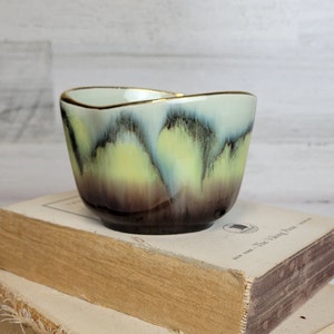 May include: A small, heart-shaped ceramic bowl with a light blue, green, and brown glaze. The bowl has a gold rim and is sitting on top of two books.