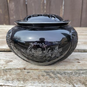 May include: A black, glossy decorative bowl with a lid. The bowl has etched floral designs and embossed patterns. The lid has small holes. The bowl is on a weathered wooden surface.