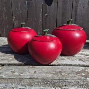May include: Three red apple-shaped canisters with removable lids. Each container features a smooth, rounded red body, a green stem handle on top, and a silver-toned leaf accent attached to the lid. The set includes three distinct sizes.