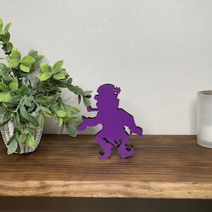 May include: Purple wooden cutout of a leprechaun with a hat and a pipe. The leprechaun is standing on a wooden shelf with a potted plant and a candle in the background.