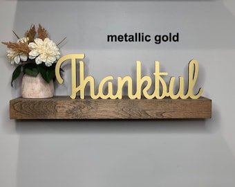 May include: A metallic gold cutout sign featuring the word Thankful in a cursive script font, positioned next to a small vase containing white flowers and dried foliage on a dark brown wooden shelf. The text metallic gold is visible.