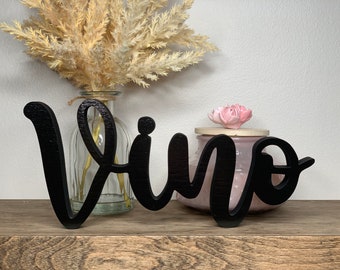 May include: Black wooden sign with the word "Vino" in cursive script. The sign is sitting on a wooden surface with a vase of dried flowers and a pink candle jar in the background.