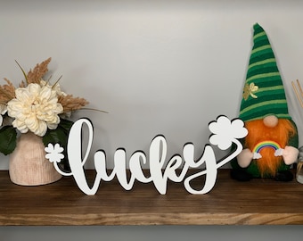 May include: White wooden sign with the word "lucky" in cursive script, with a small shamrock cutout at the end. The sign is on a wooden shelf with a gnome holding a rainbow and a vase of white flowers.