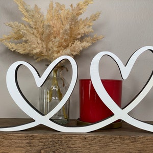 May include: Two white wooden hearts intertwined, creating a decorative piece for a home or wedding. The hearts are on a wooden shelf with a vase and a red candle holder.