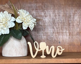 May include: A wooden sign that spells "Vino" in a cursive font, with a wine glass incorporated into the "V". The sign is light-colored and sits on a wooden shelf next to a vase of white flowers.