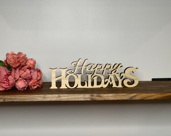 May include: Wooden sign with the text "Happy Holidays" in a decorative font. The sign is light brown and is sitting on a wooden shelf. There is a bouquet of pink peonies in the foreground.