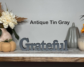 May include: A gray wooden sign that spells out the word "Grateful" in a decorative font. The sign is painted in an antique tin gray color and is displayed on a wooden shelf with decorative pumpkins and flowers.