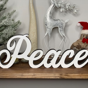 May include: White wooden sign with the word "Peace" in a cursive font. The sign is sitting on a wooden shelf with a small Christmas tree, a silver reindeer figurine, and a brown bird figurine wearing a Santa hat in the background.