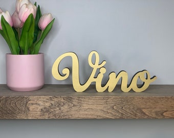 May include: Gold-painted wooden sign with the word "Vino" in a cursive font. The sign is sitting on a wooden shelf.